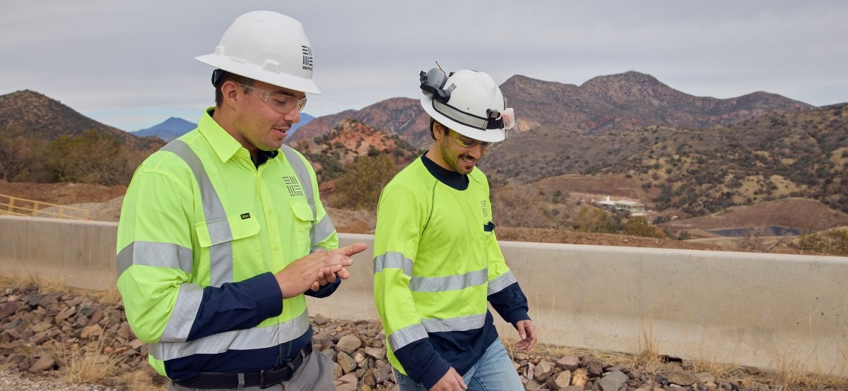 Two South32 workers in high-visibility clothing and hard hats walk together at the Hermosa Project, with the Arizona hills behind them.
