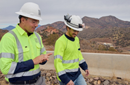 Two South32 workers in high-visibility clothing and hard hats walk together at the Hermosa Project, with the Arizona hills behind them.